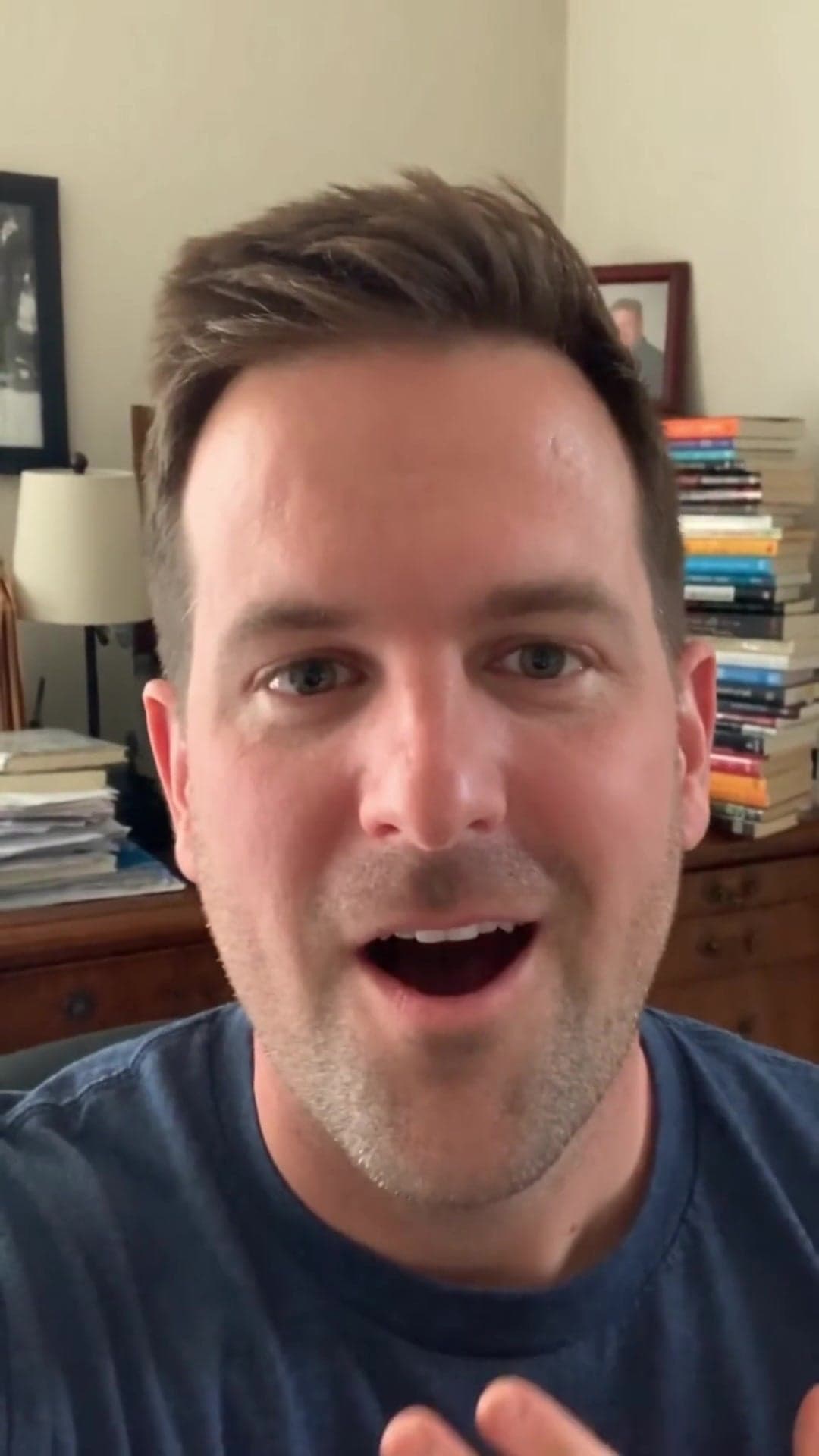 Man in a navy shirt speaking to the camera with books behind him